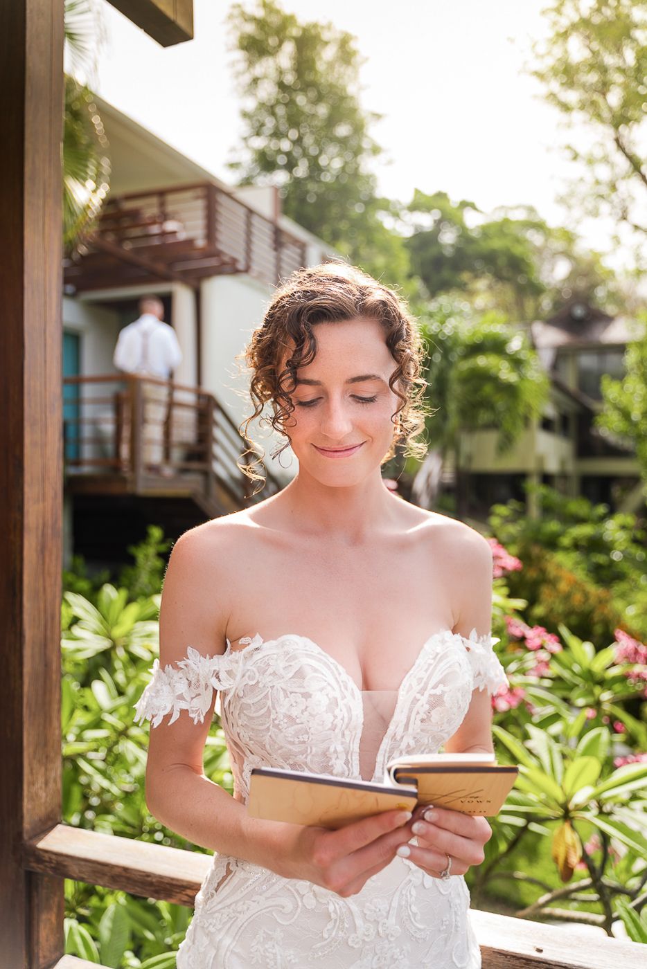 A bride reads from a book while sitting in an outdoor tropical garden setting.