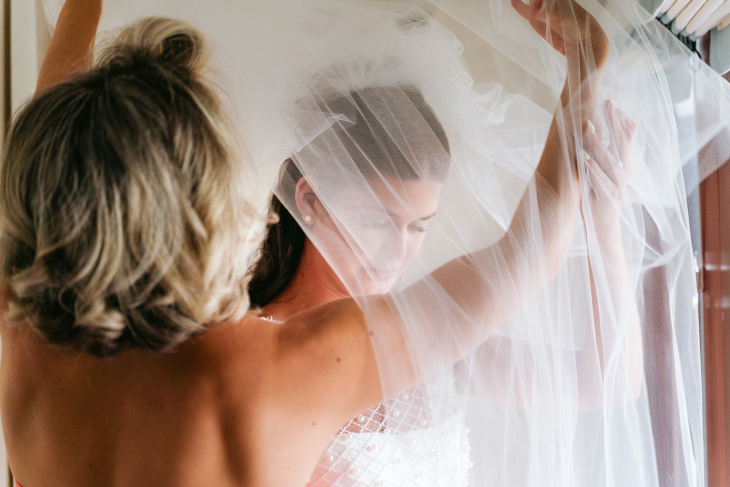 A person helps adjust a delicate, flowing white wedding veil in soft natural lighting.