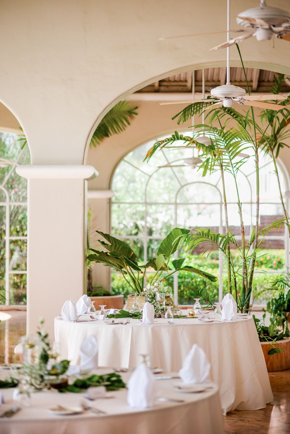 Tropical indoor dining setup with white tablecloths and palm plants under arched columns.