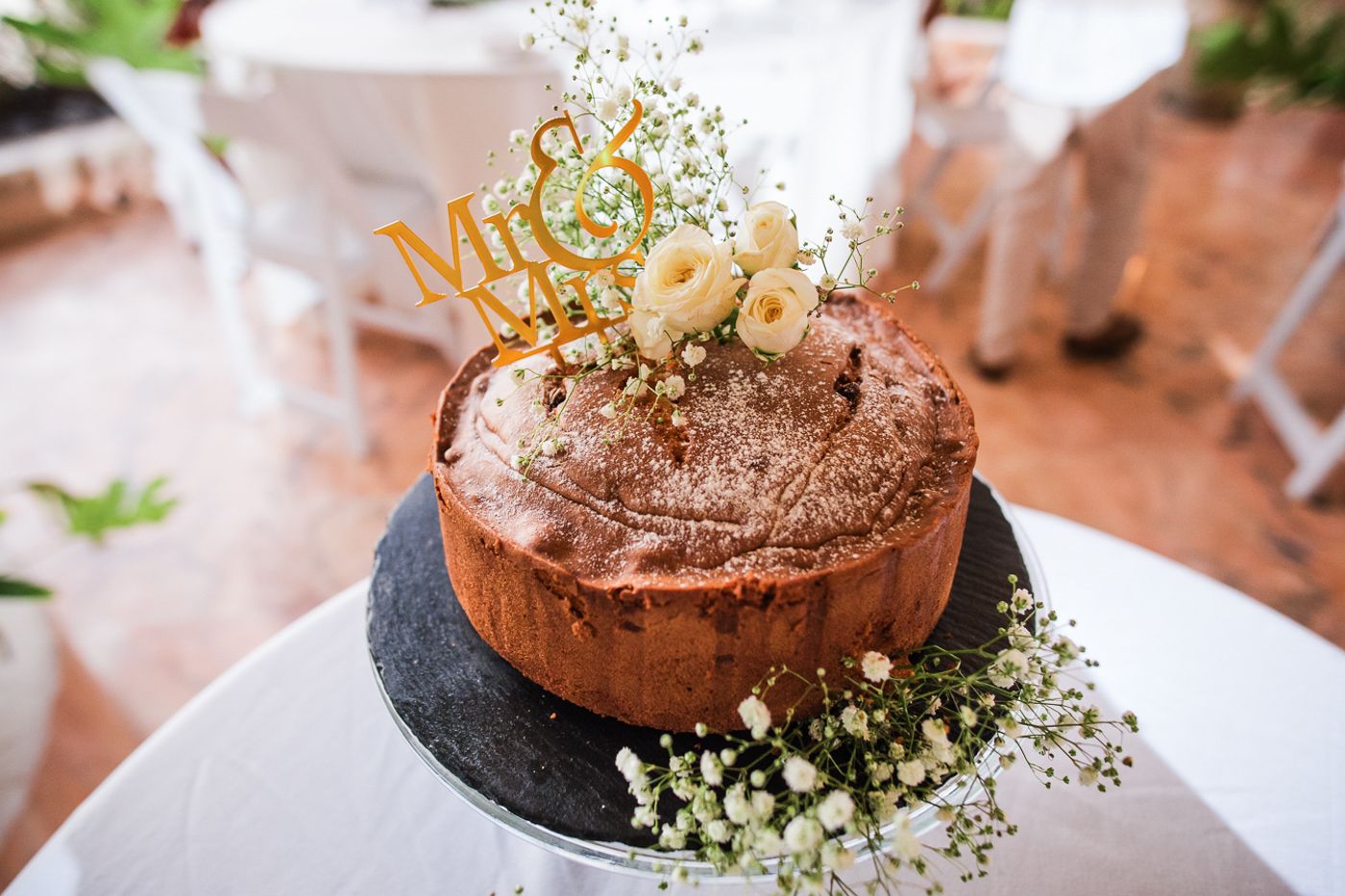 Chocolate wedding cake decorated with gold accents and baby's breath flowers on a white tablecloth.