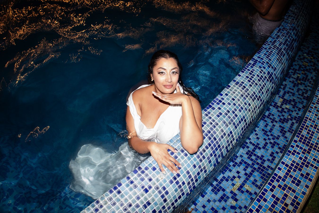 Someone lounges along the edge of a pool with decorative mosaic tiles at night.
