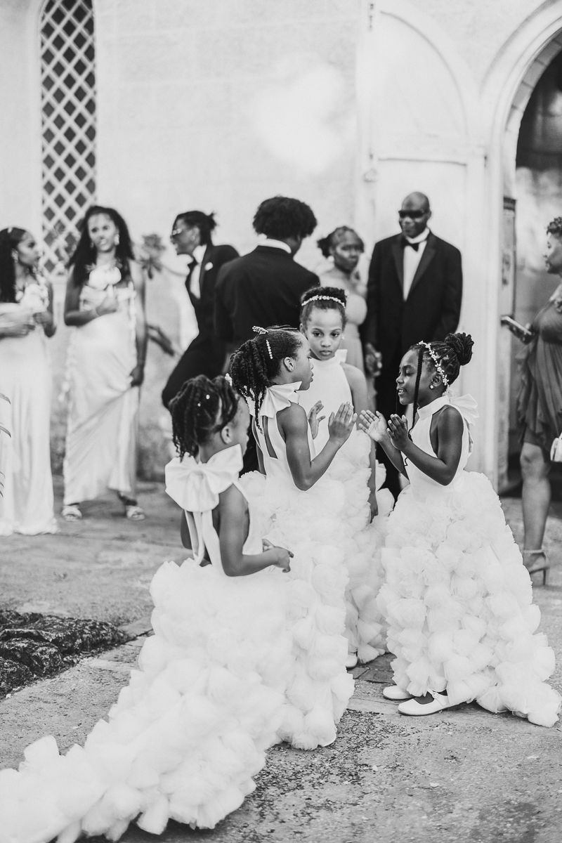 Black and white wedding photo showing people gathered outdoors with elegant white dresses and formal attire.
