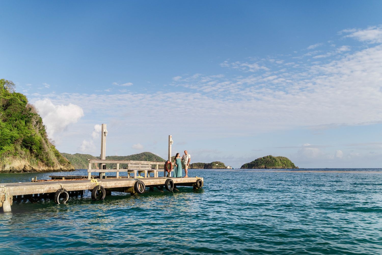 Two people stand on a wooden dock overlooking turquoise Caribbean waters with lush green islands in the distance.