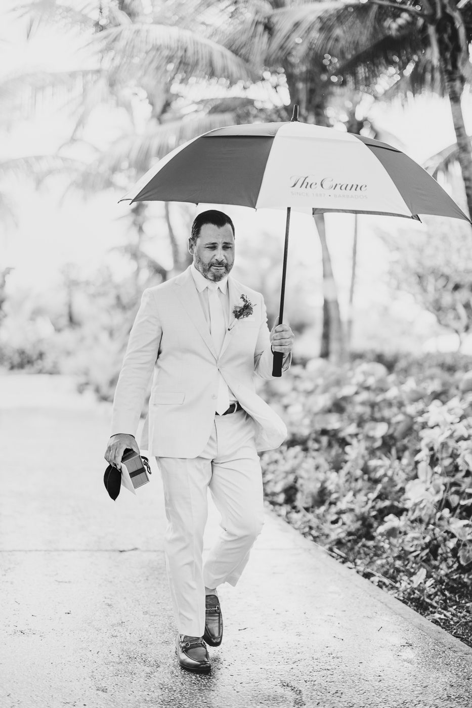 Groom in white suit walks with umbrella labeled The Crane, carrying shoes on a rainy tropical wedding day.