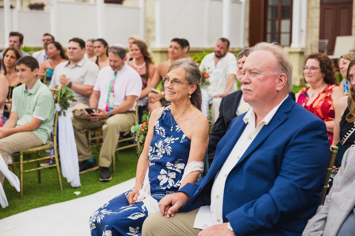 Guests seated outdoors at a wedding ceremony, including a woman in a floral dress and a man in a navy suit.