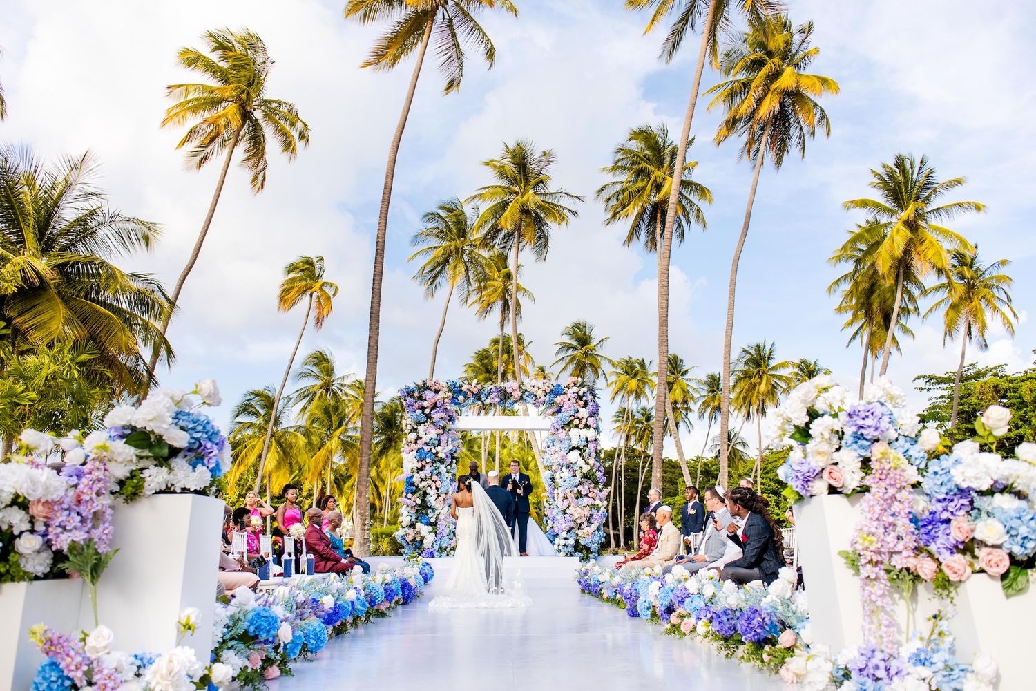 Tropical beach wedding ceremony with blue and purple floral arch, white aisle, and tall palm trees under a bright sky.