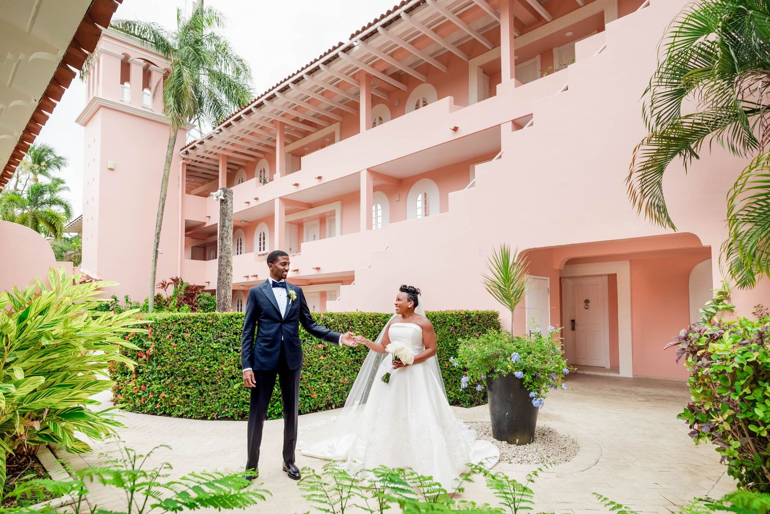 Bride and groom hold hands in lush garden courtyard of pink resort building on their wedding day.