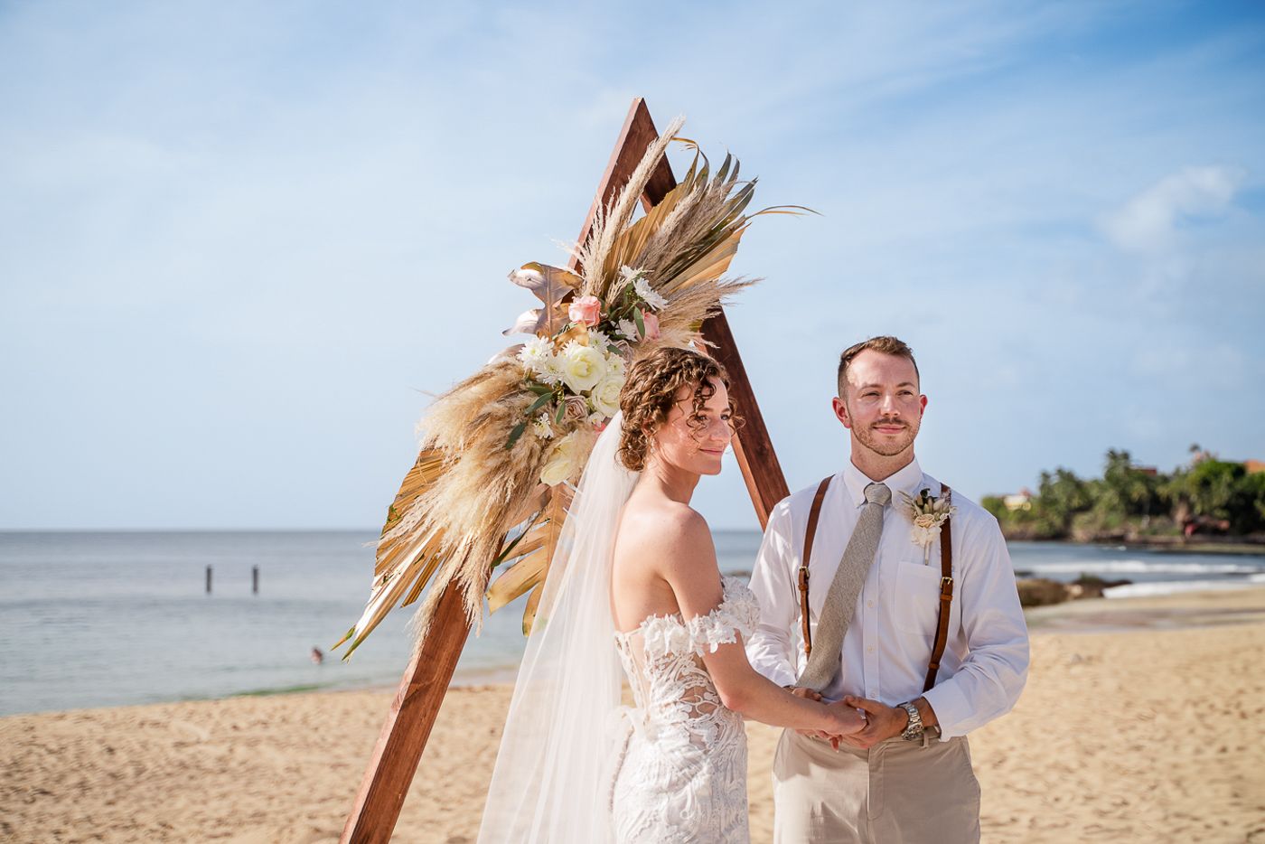 Beachfront wedding ceremony with flowing white dress and dried floral bouquet against blue sky.