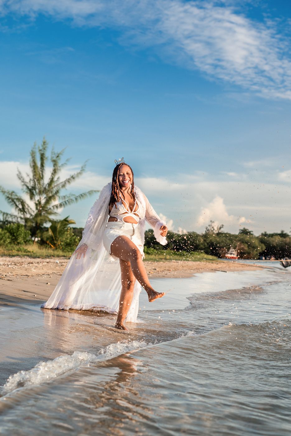 Joyful bride splashing in ocean waves while wearing her wedding dress on a tropical beach.