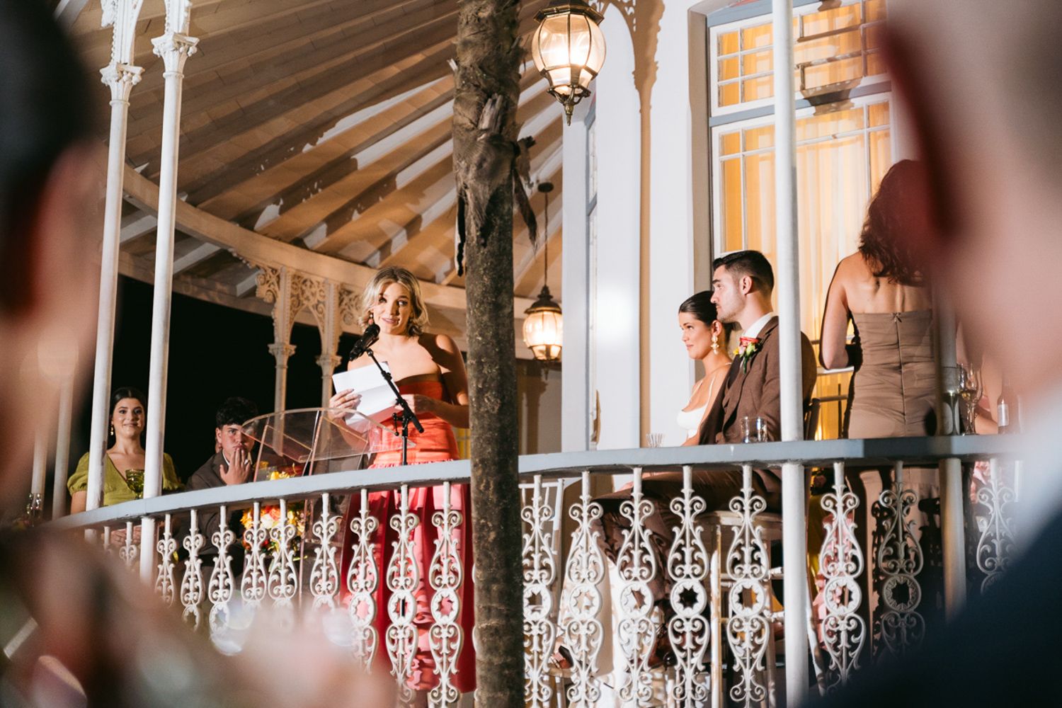 A group mingles on an elegant balcony with decorative white railings and warm lighting in a vintage-style setting.
