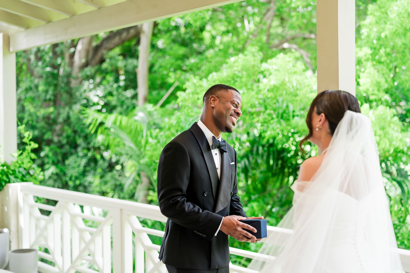 A covered outdoor porch with white railings and tropical landscaping sets the scene for a wedding moment.
