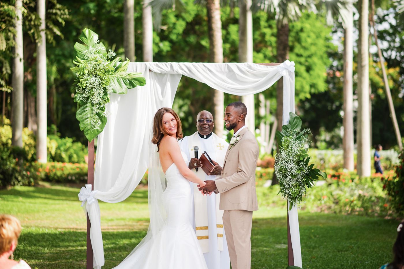 Wedding ceremony under white fabric arch decorated with greenery in tropical garden setting.