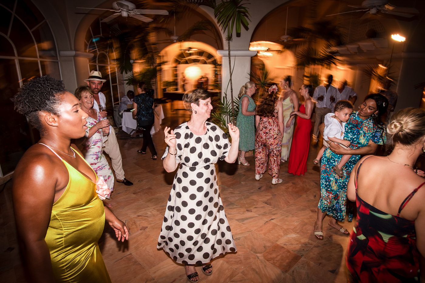 Guests dancing at a tropical-themed wedding reception in an elegant venue with arched windows and palm trees.