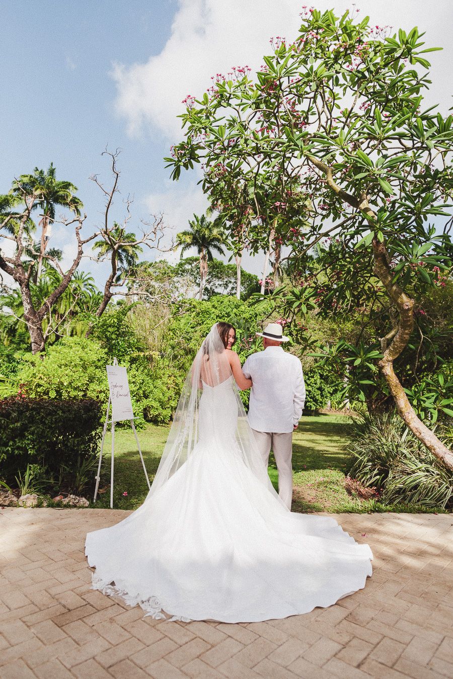 A couple in wedding attire walks along a brick pathway surrounded by lush tropical gardens and palm trees. Fairmont Barbados
