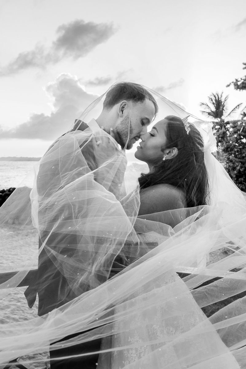 Black and white beach wedding photo of bride and groom almost kissing under flowing veil at sunset.