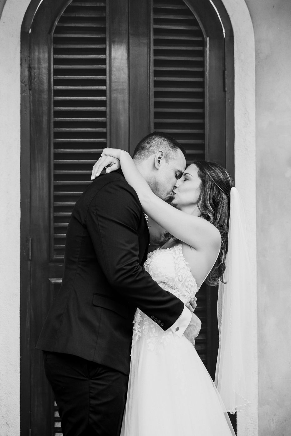 Black and white photo of a bride and groom sharing a romantic kiss in front of elegant shuttered doors.