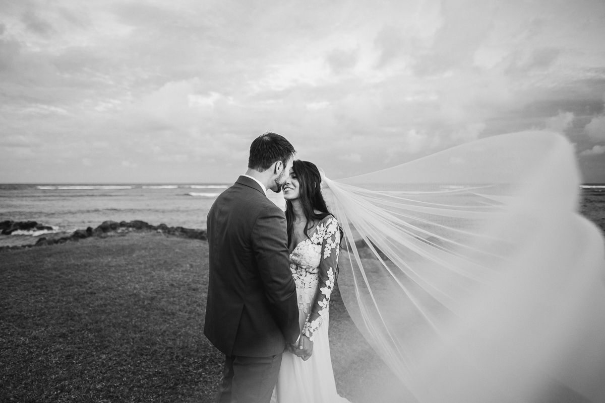 Black and white photo of bride and groom embracing near ocean, veil flowing in wind.