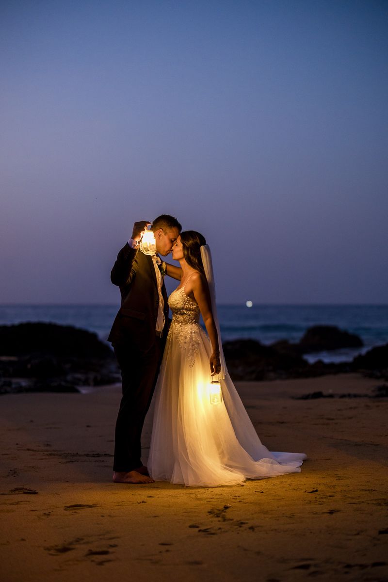 Bride and groom kissing on a beach at dusk, holding glowing lanterns, with ocean waves in the background.