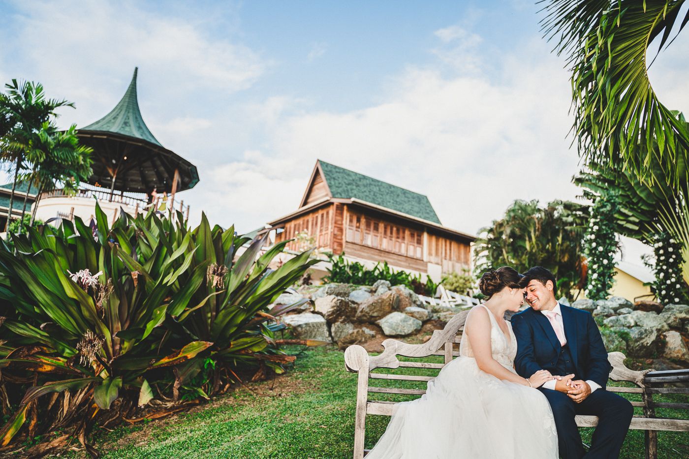 Bride and groom share a romantic kiss in a lush tropical garden with a wooden pavilion backdrop.