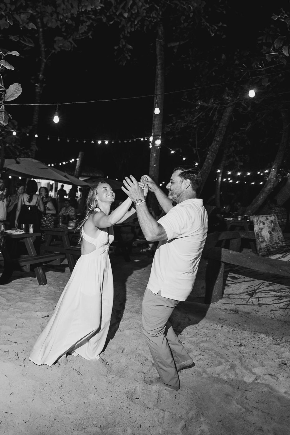 Couple dancing on sandy beach at night under string lights in black and white wedding photo.
