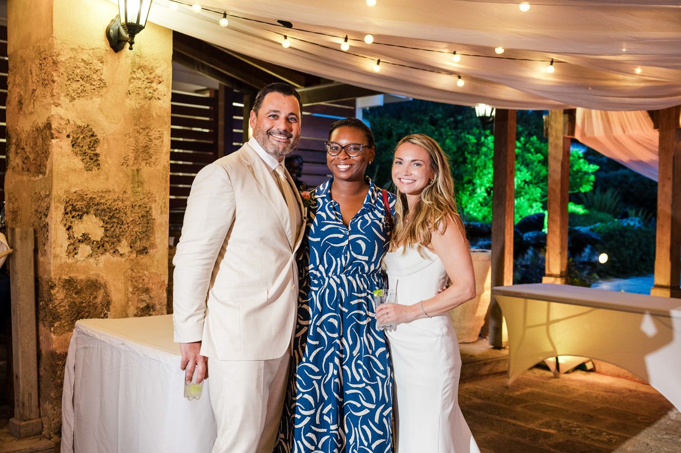 Three people smiling together at an outdoor evening event, two in white and one in a blue patterned dress.