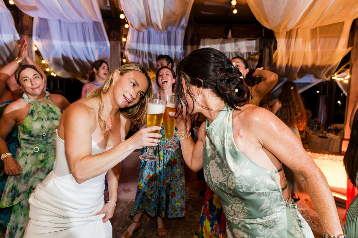 Bride in white dress toasting beer glasses with guest in green dress on wedding reception dance floor.