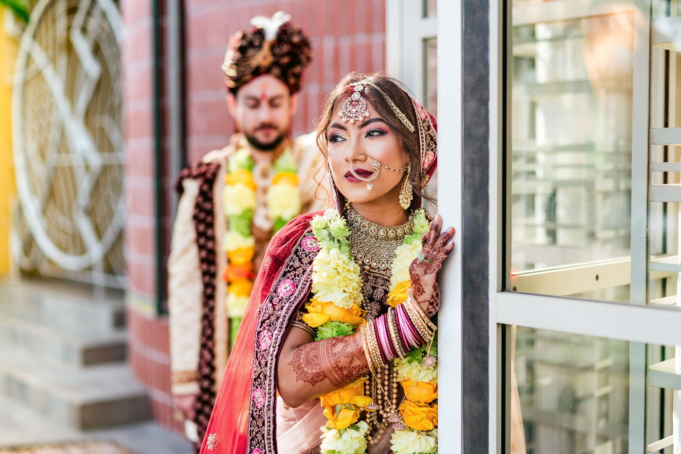 Indian bride and groom in traditional wedding attire with floral garlands posing at a doorway.