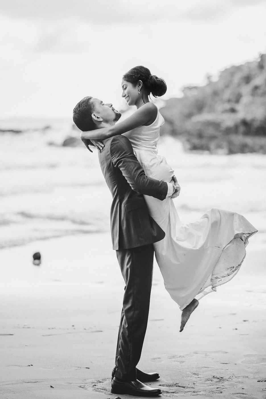 Groom lifting bride in white dress on beach in romantic black and white wedding photo.