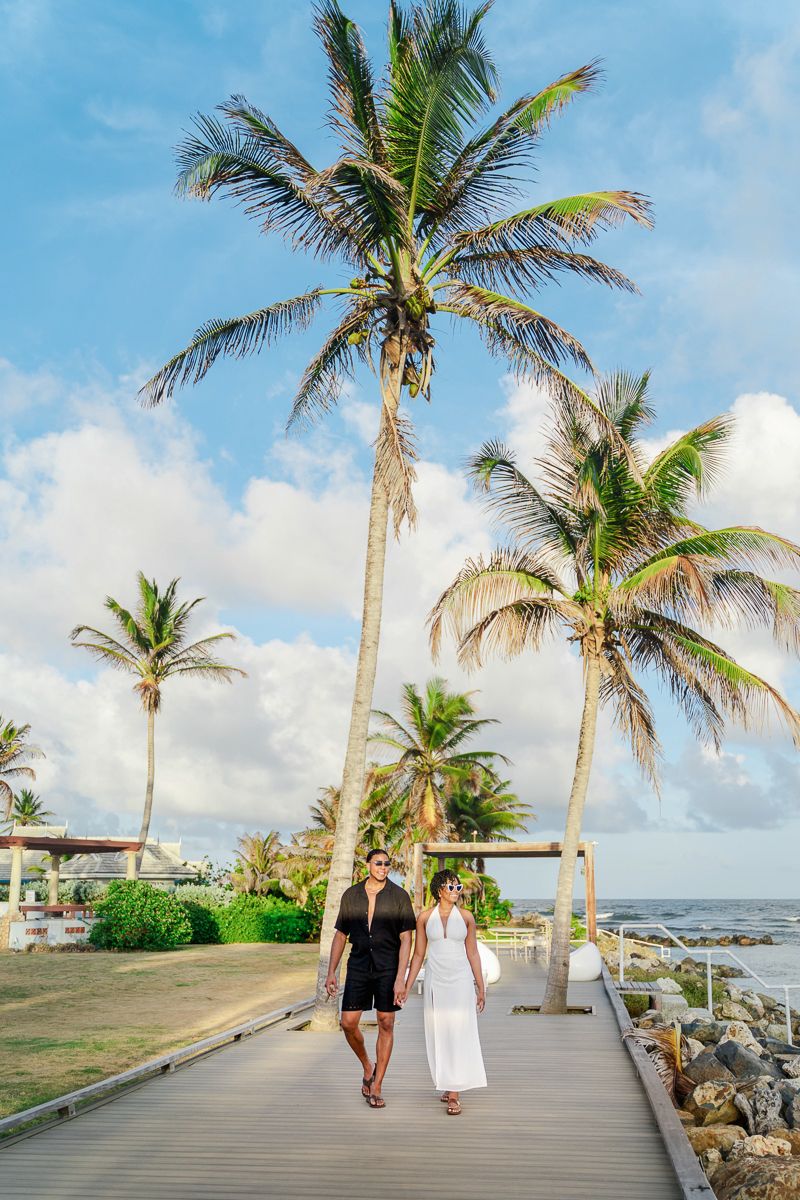 Couple walking on a tropical boardwalk lined with tall palm trees near the ocean under a partly cloudy blue sky.