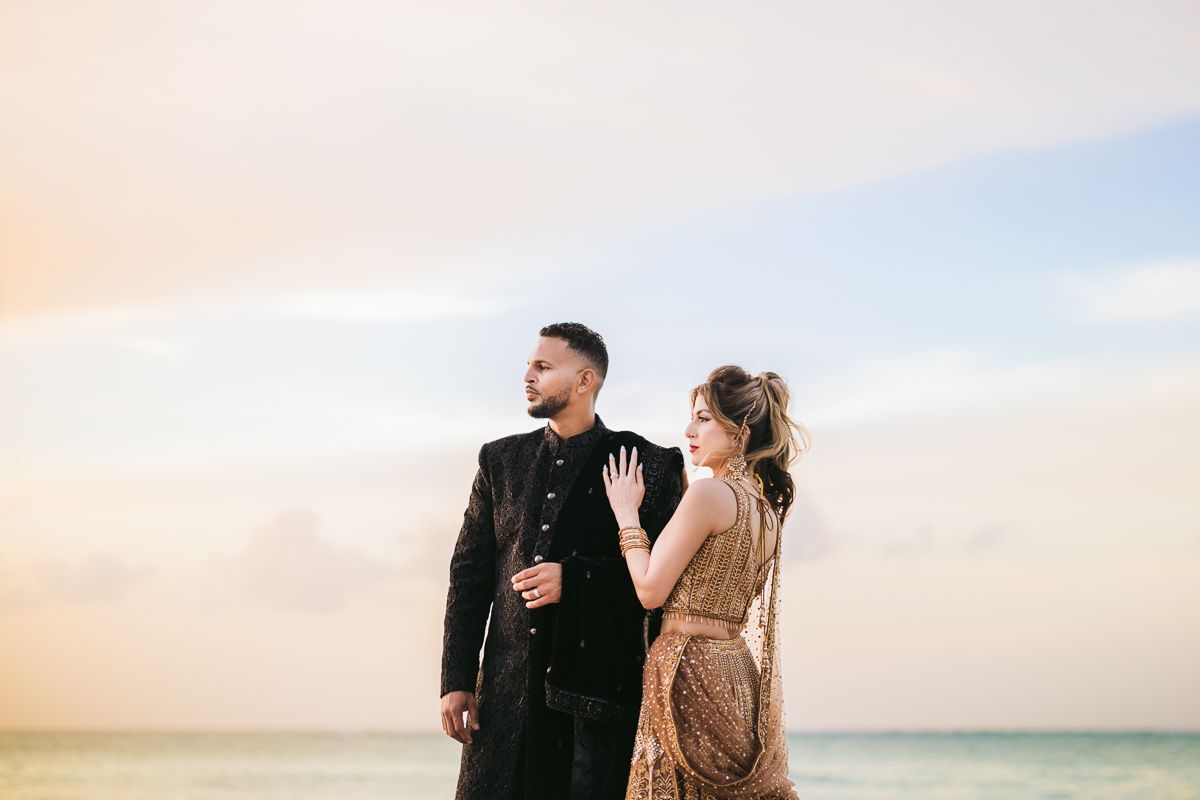 Couple in traditional South Asian wedding attire poses on a beach at sunset, woman in gold sari, man in black sherwani.