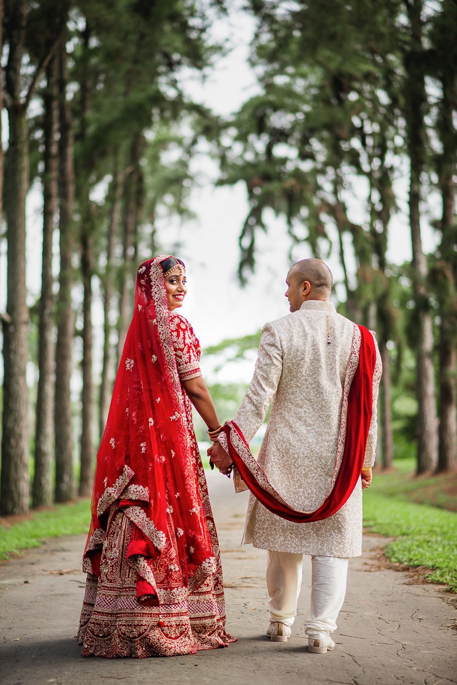 Indian bride in red lehenga and groom in white sherwani hold hands walking through tree-lined path on wedding day.