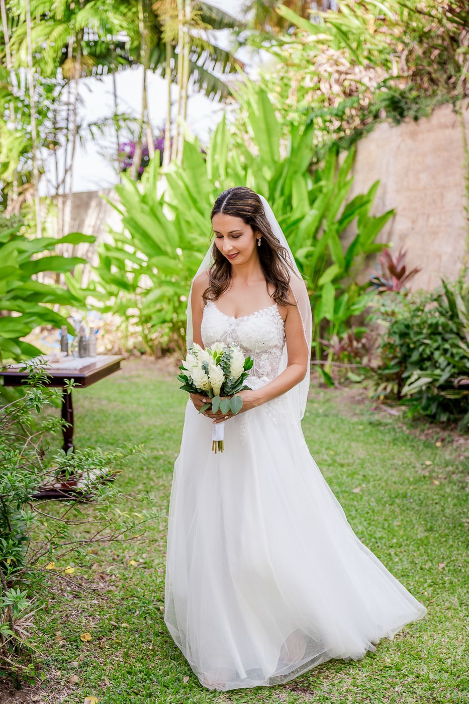 Bride in white lace A-line gown holding white bouquet, standing in lush tropical garden on wedding day.