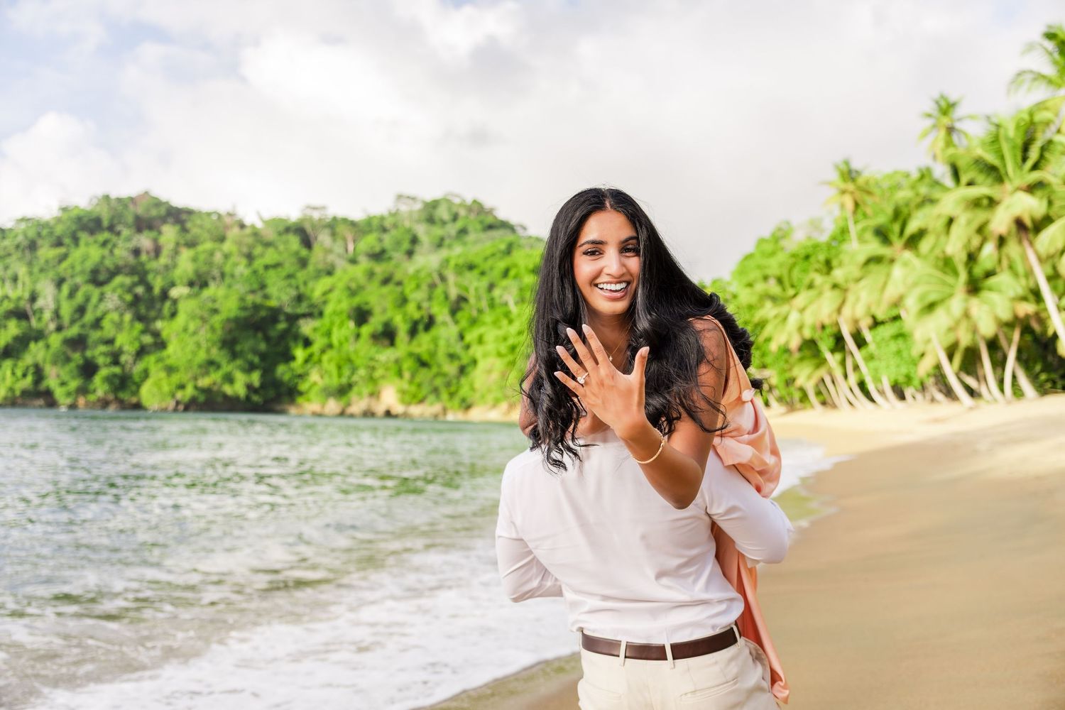 Woman showing engagement ring on tropical beach with palm trees and turquoise water in background.