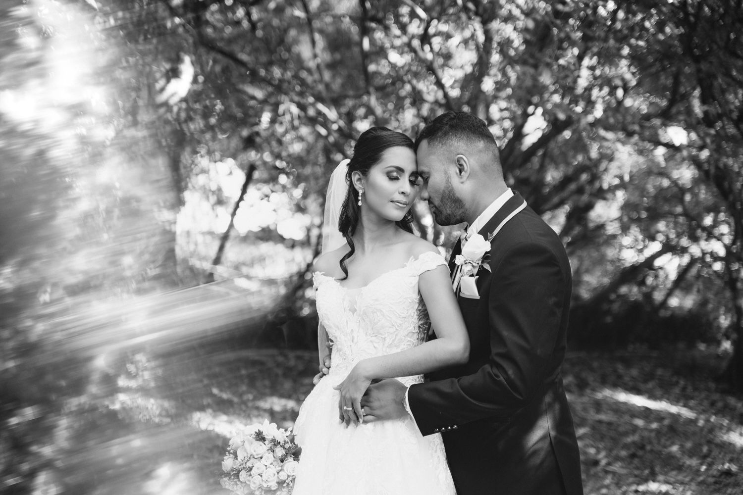 Black and white photo of a bride and groom sharing an intimate moment surrounded by trees on their wedding day.