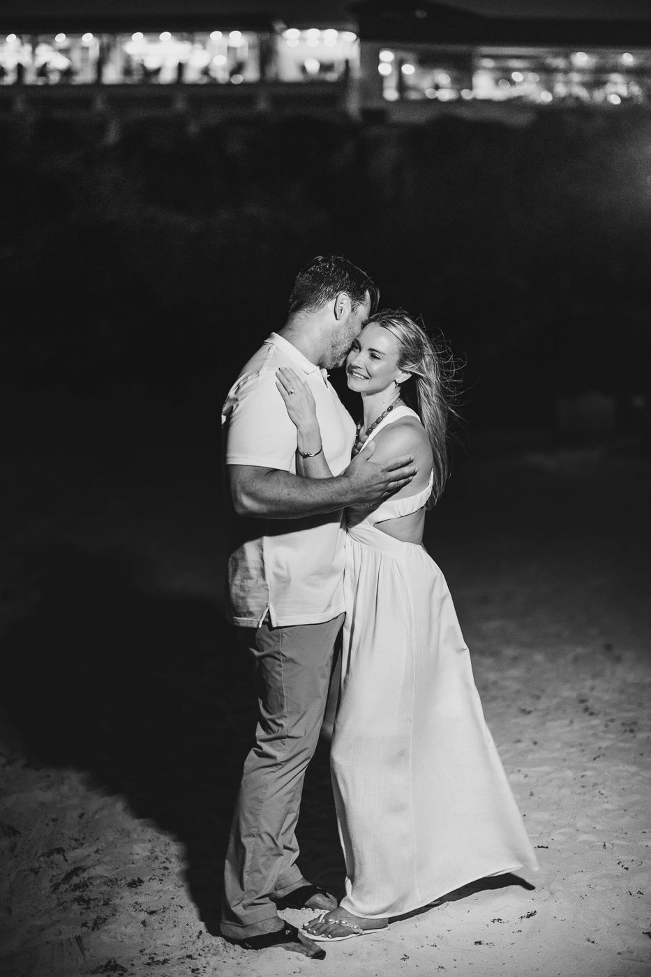 Black and white photo of a couple embracing and dancing on a beach at night with festive lights in the background.