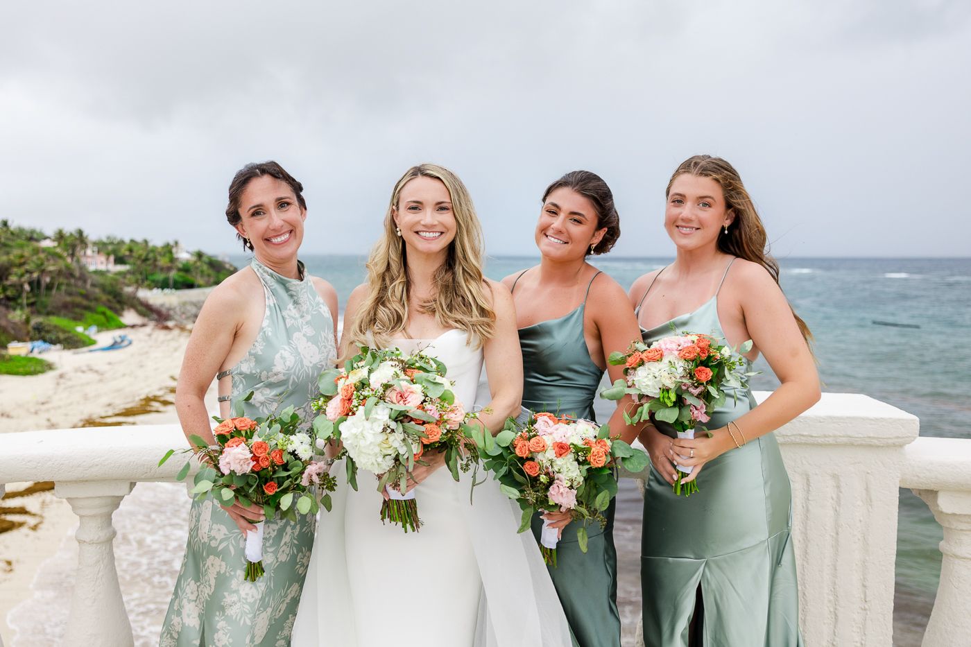 Bride in white gown with three bridesmaids in sage green dresses holding coral and white bouquets at oceanside wedding venue.