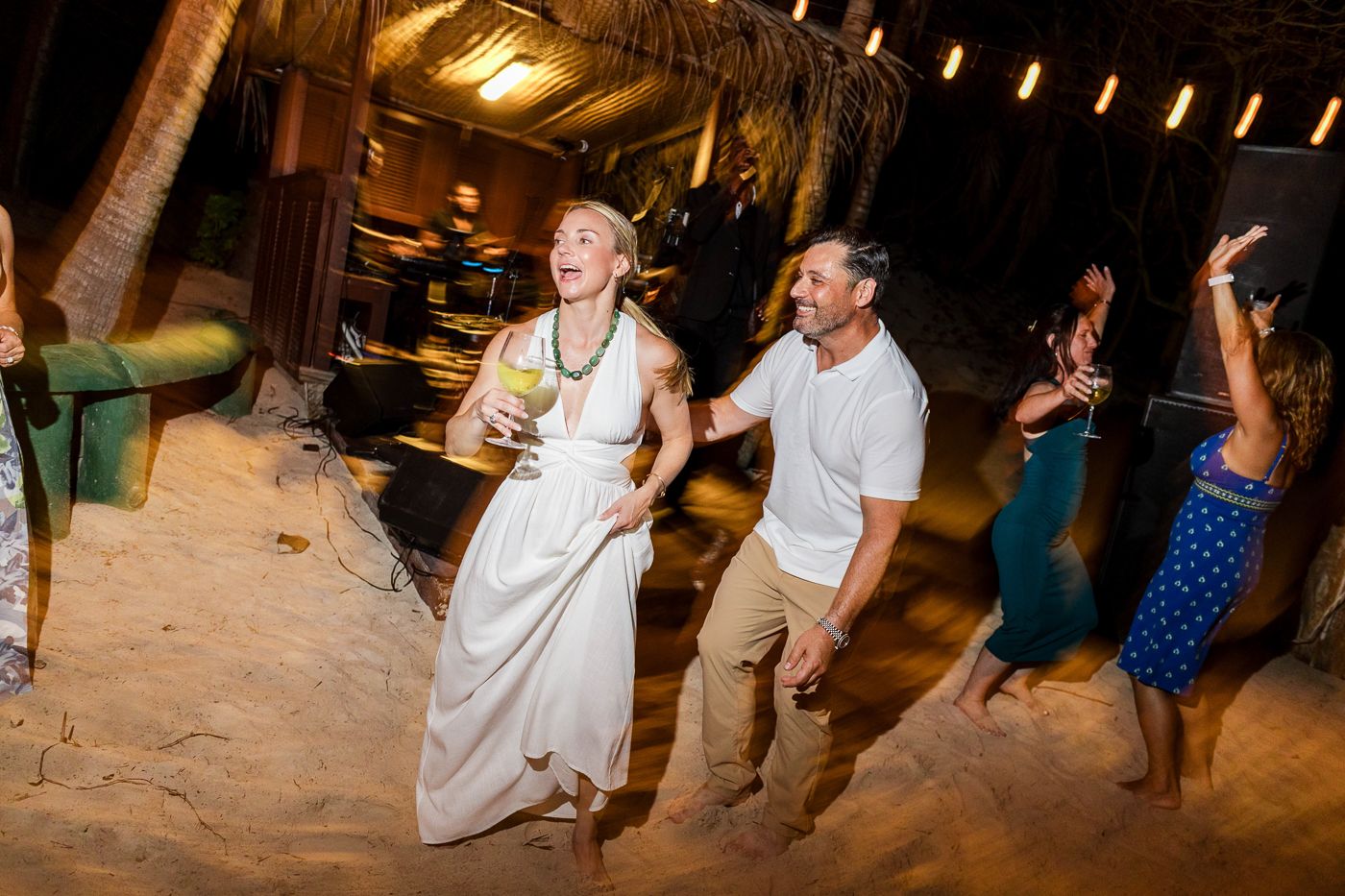 Couple dancing joyfully on sandy beach at night during outdoor wedding reception with tropical palm trees.