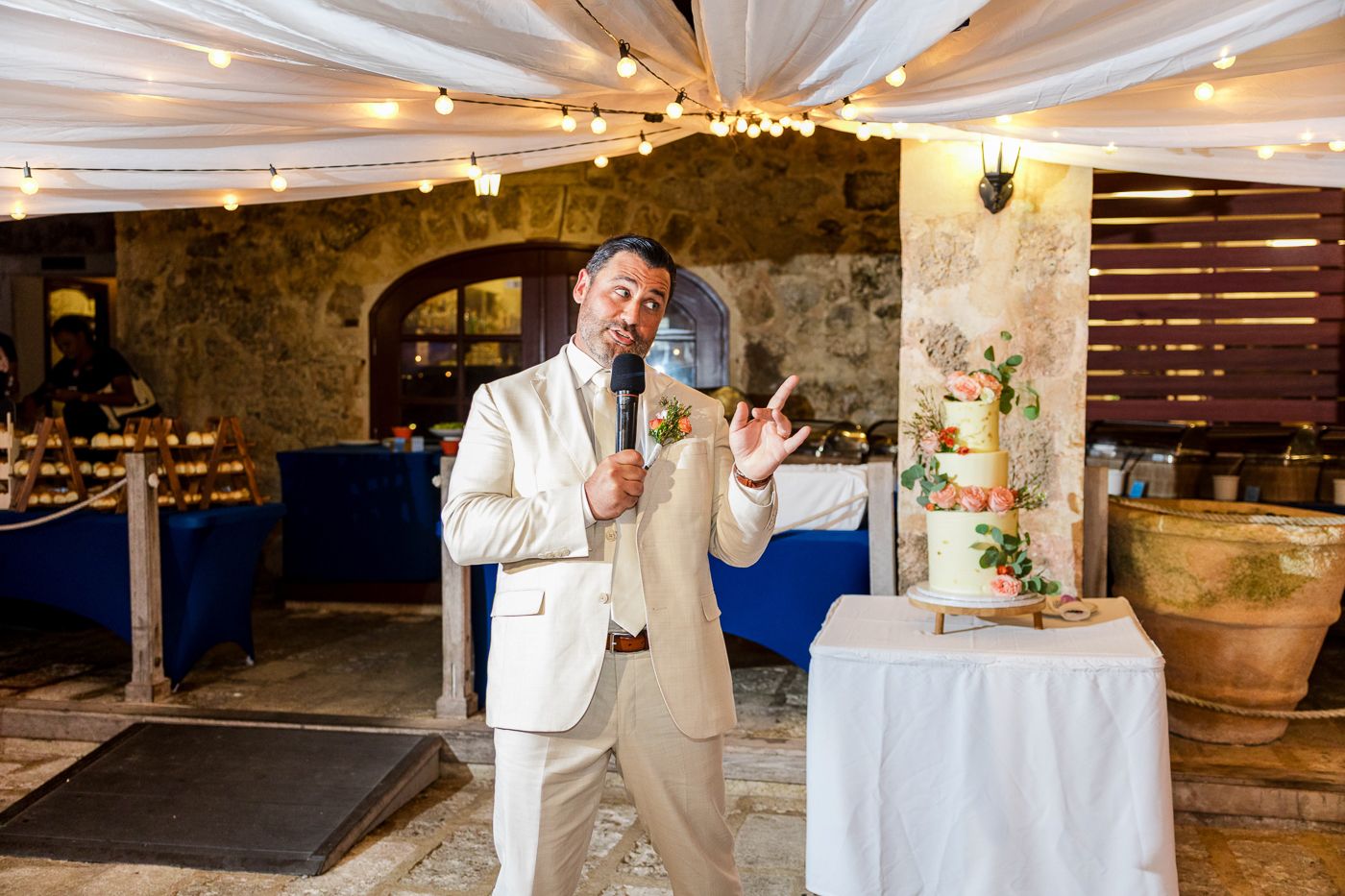 Man in white blazer and tan pants smiling and gesturing near a wedding cake at an elegant stone venue.