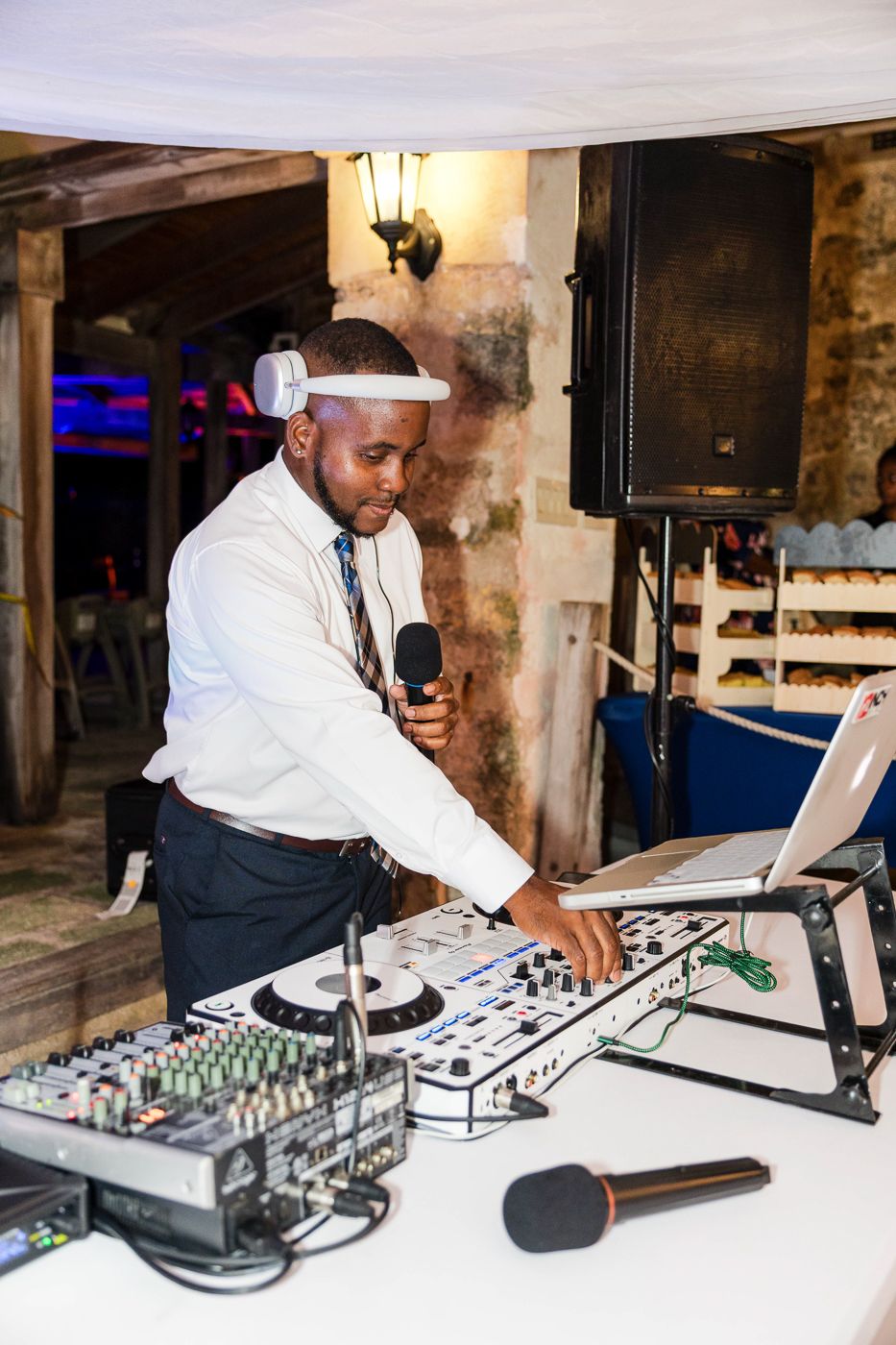 A DJ in a white shirt and cap operates a mixing console while holding a microphone at an indoor event.