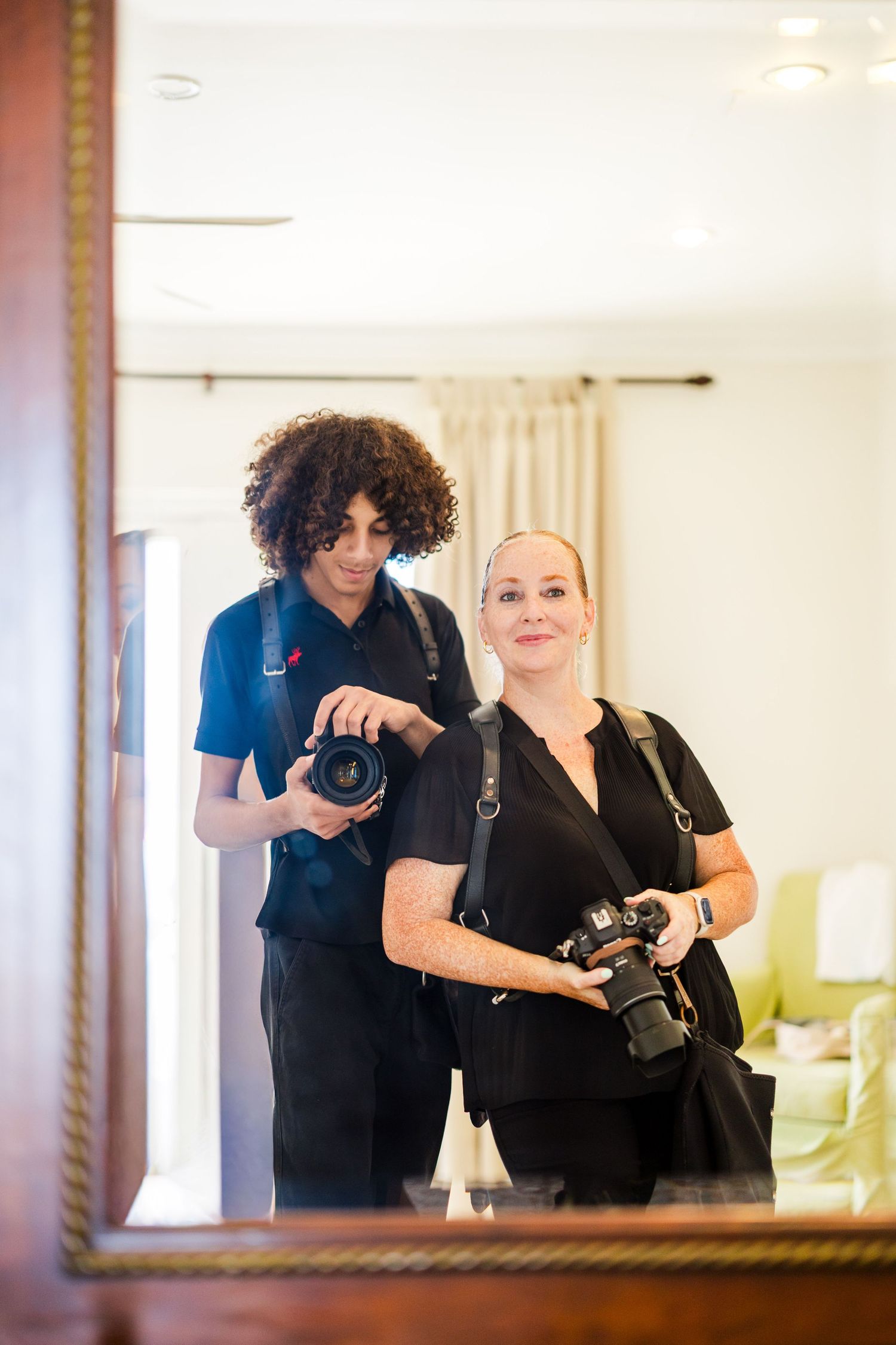 Two photographers in black outfits take a mirror selfie while holding DSLR cameras in a bright, modern interior.