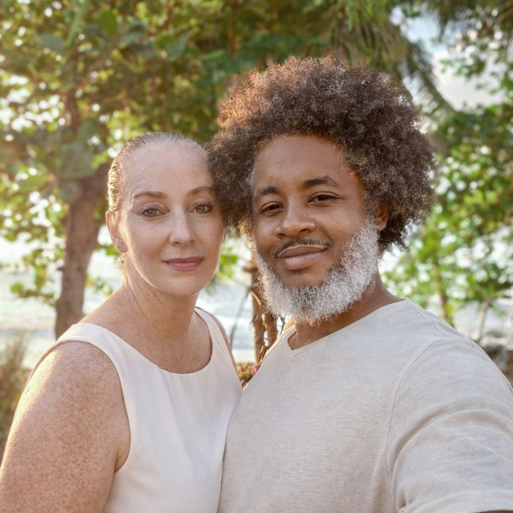 A smiling interracial couple poses outdoors near green trees, woman in white top and man with curly hair and beard.