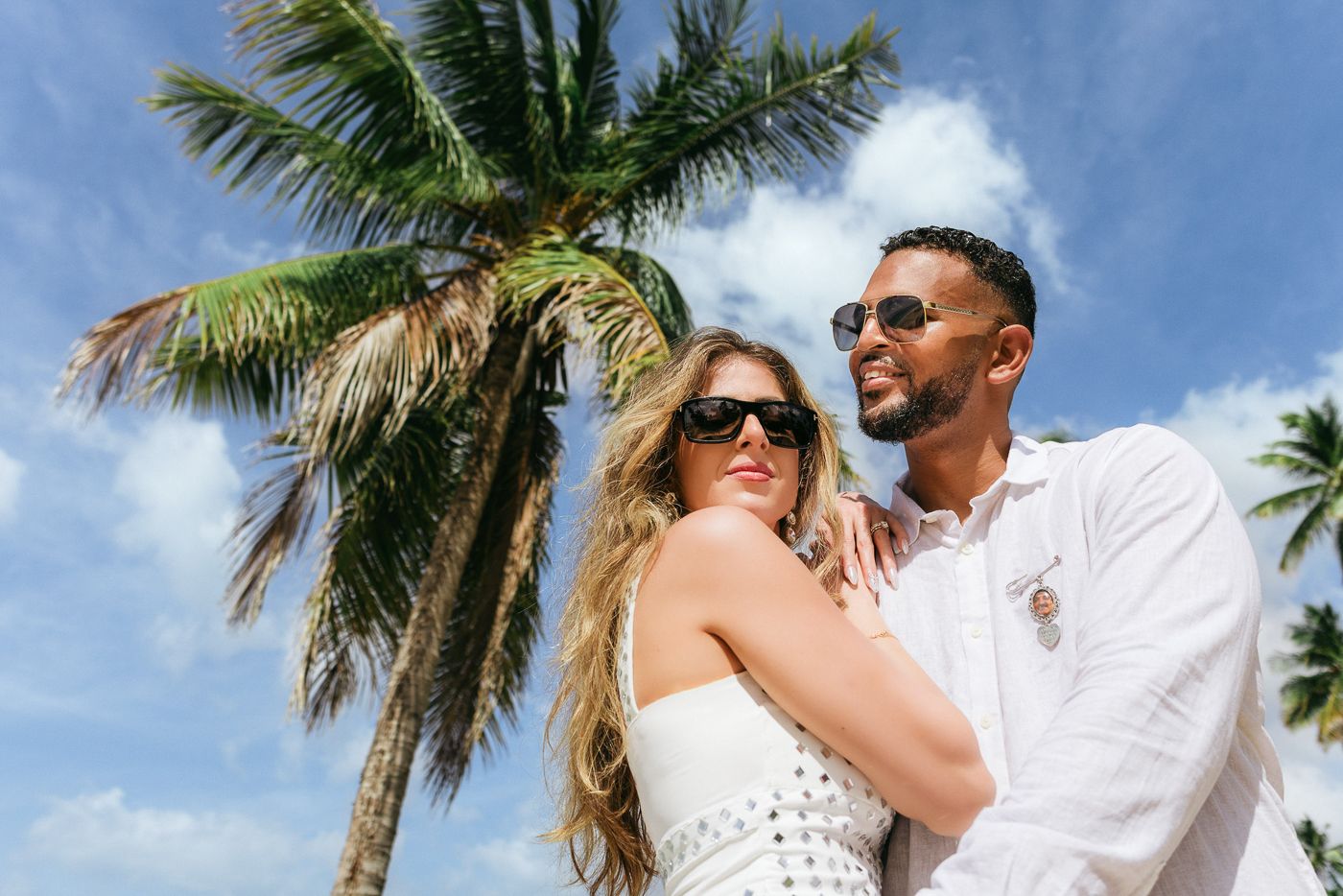 A couple in white outfits embraces and kisses on a tropical beach with palm trees and blue sky in the background.