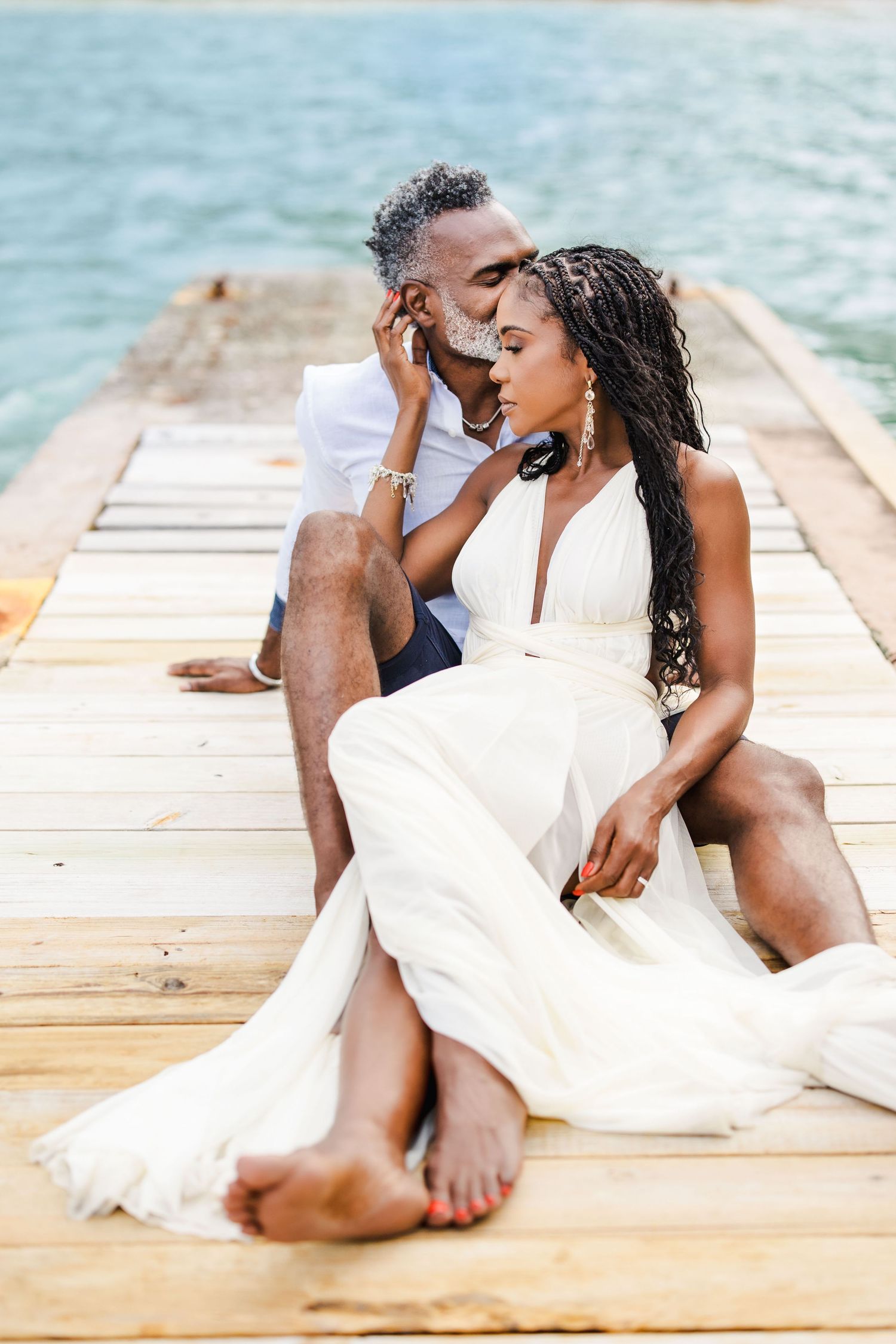 Romantic couple in white attire sharing intimate moments on a wooden dock by the water.