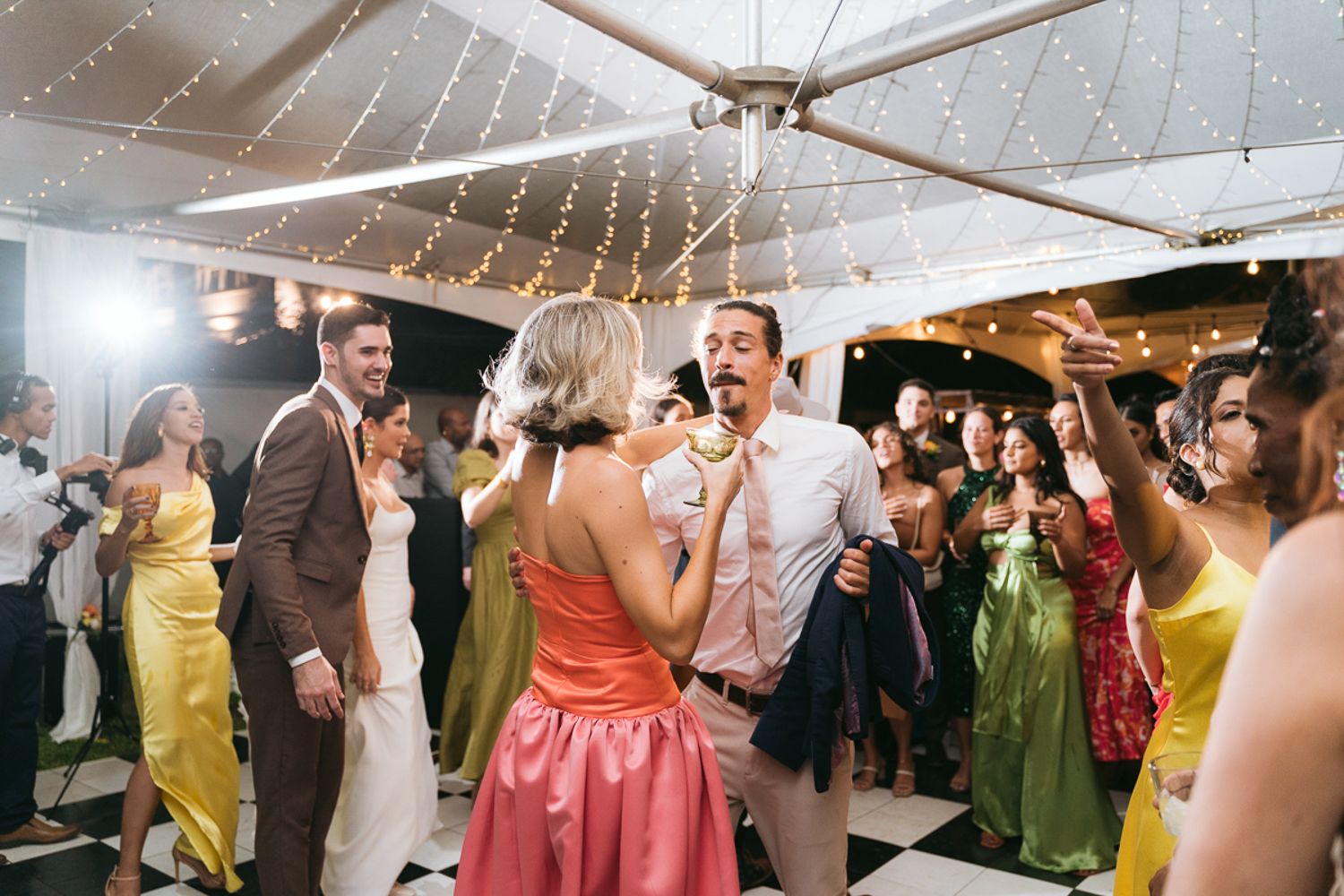 Guests dance under twinkling string lights at a wedding reception in a bright indoor venue.