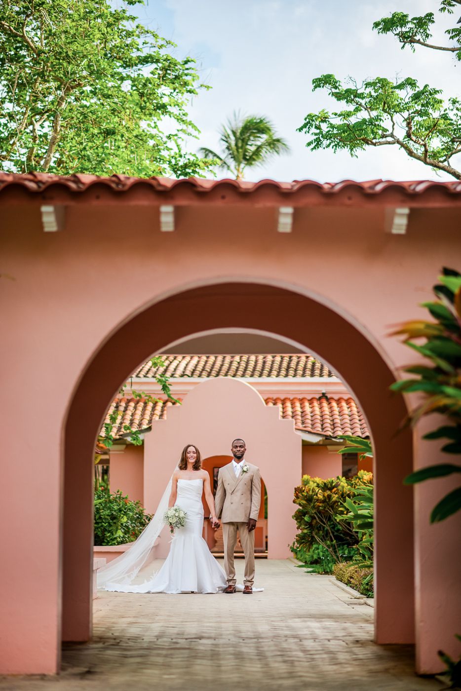 Wedding couple in tropical setting framed by pink Spanish arches and lush greenery.