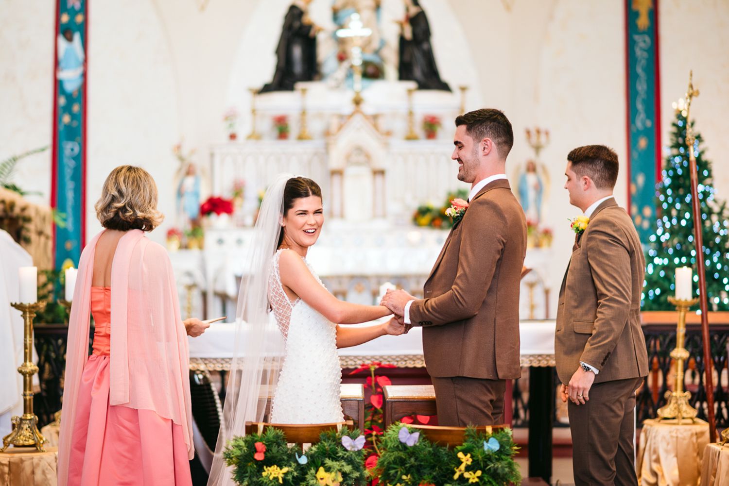 Wedding ceremony in a bright church with pink and white floral decorations and pews filled with guests.