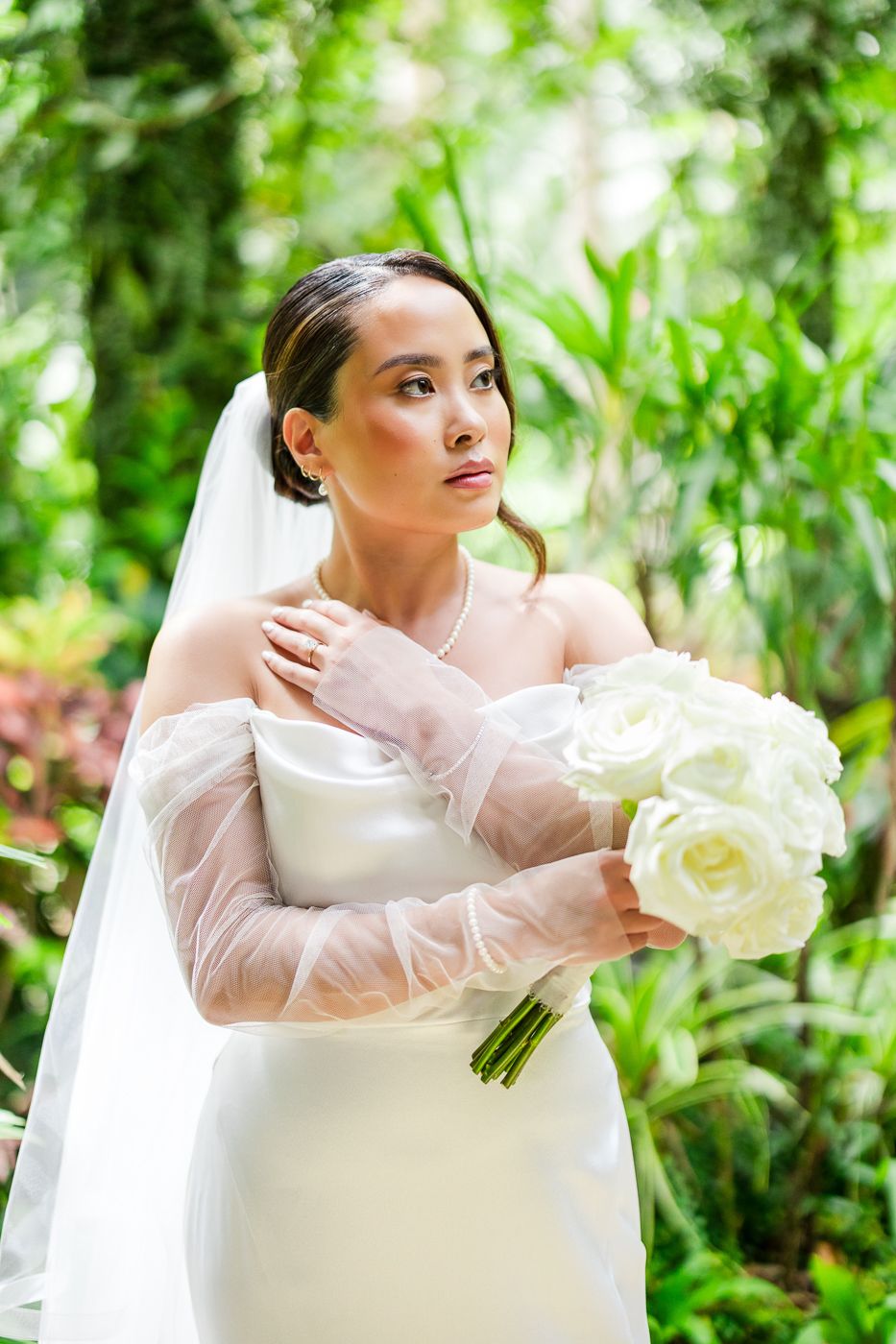 Elegant bride in a white gown with sheer sleeves holds a bouquet of white roses against a lush garden backdrop.