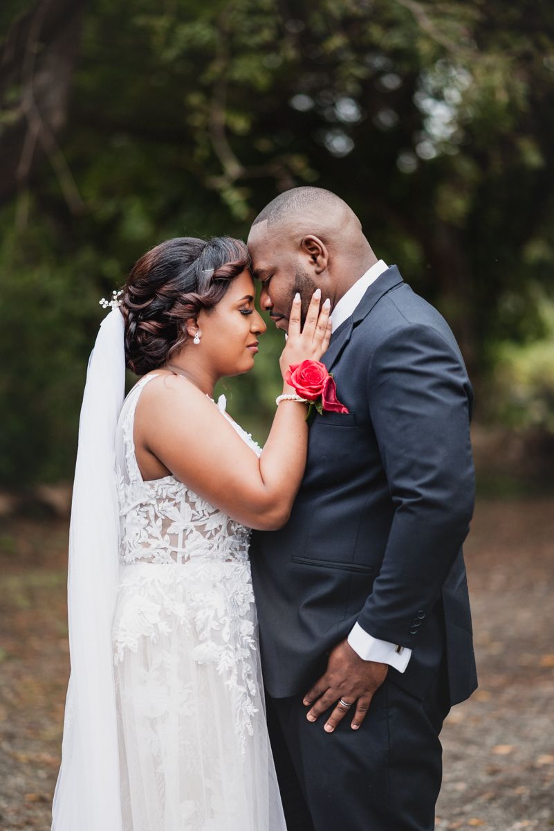 A bride and groom share an intimate forehead touch moment outdoors, she in lace gown with veil, he in dark suit with red rose.