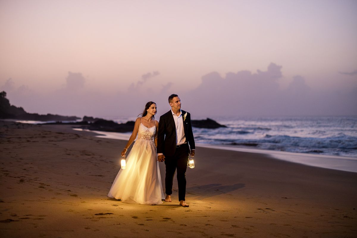 Bride and groom walk hand in hand on a beach at dusk, holding lanterns with ocean waves in the background.