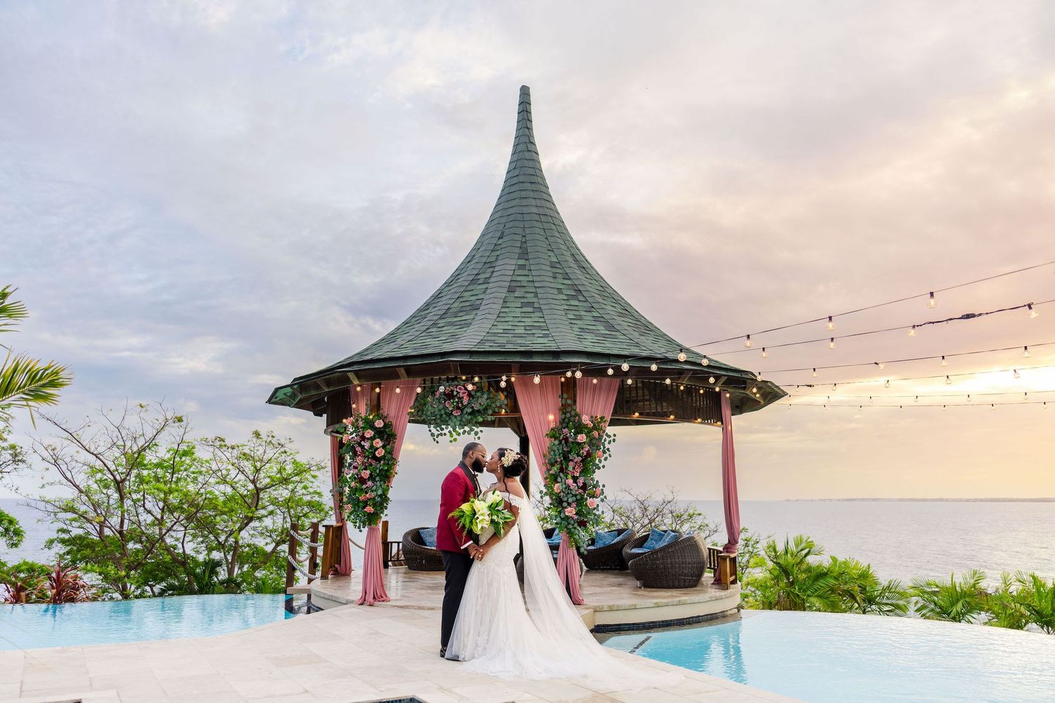 Couple kissing under elegant gazebo with ocean view at sunset during tropical destination wedding ceremony.